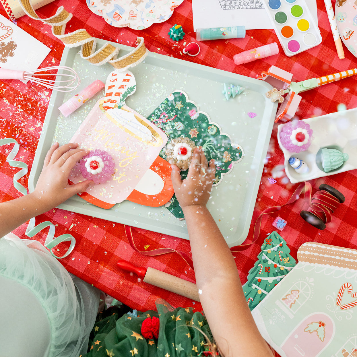 Children's hands playing with Christmas-themed toys on a red tablecloth.