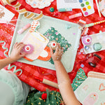Children's hands playing with Christmas-themed toys on a red tablecloth.